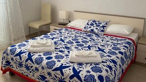 Een net opgemaakt bed met een blauw-wit gebloemd sprei, een wit kussen en twee opgevouwen handdoeken op het bed. Er staat een stoel en een nachtkastje met een lamp naast het bed.