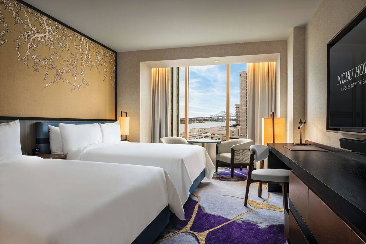 A hotel room with two beds, a desk, a chair, a TV, and a view of the city.