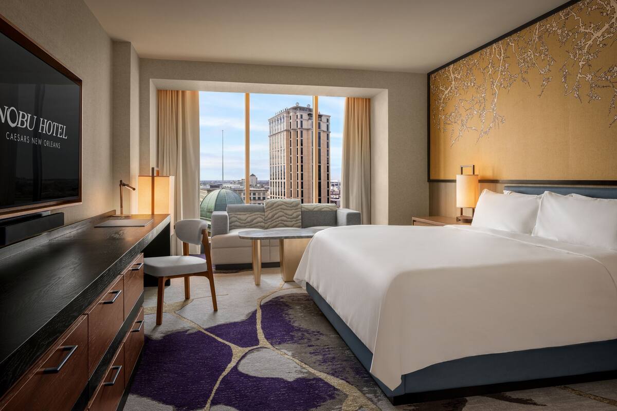 A hotel room with a bed, a desk, a chair, a TV, and a view of the city.
