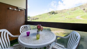 Ein Balkon mit rundem Tisch, weißen Stühlen, einem Blumentopf und Blick auf eine grüne Landschaft mit Hügeln und Windkraftanlagen.