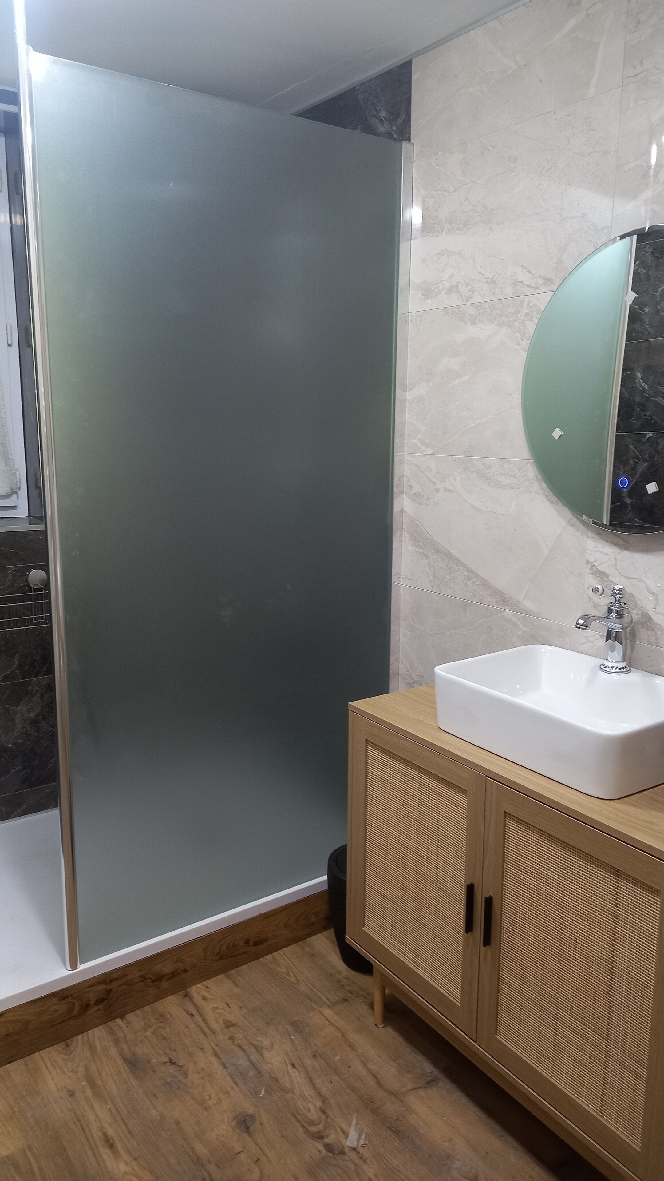 A modern bathroom with a glass shower enclosure, a wooden vanity with a sink, and a marble-tiled wall.