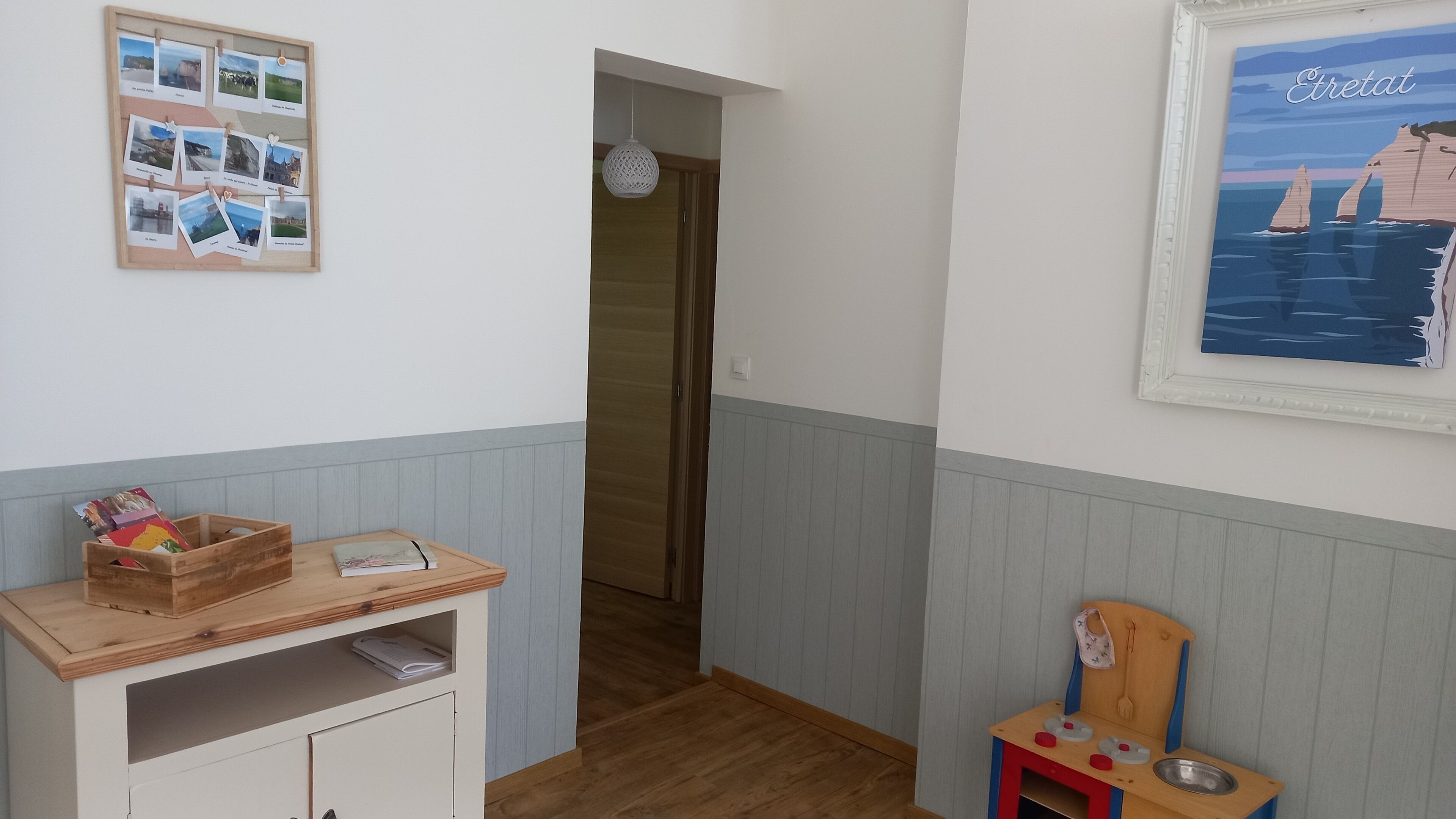 Un pasillo con piso de madera, una pequeña mesa de madera con una cesta y un libro, un collage de fotos enmarcadas en la pared, y una pintura de una escena costera.