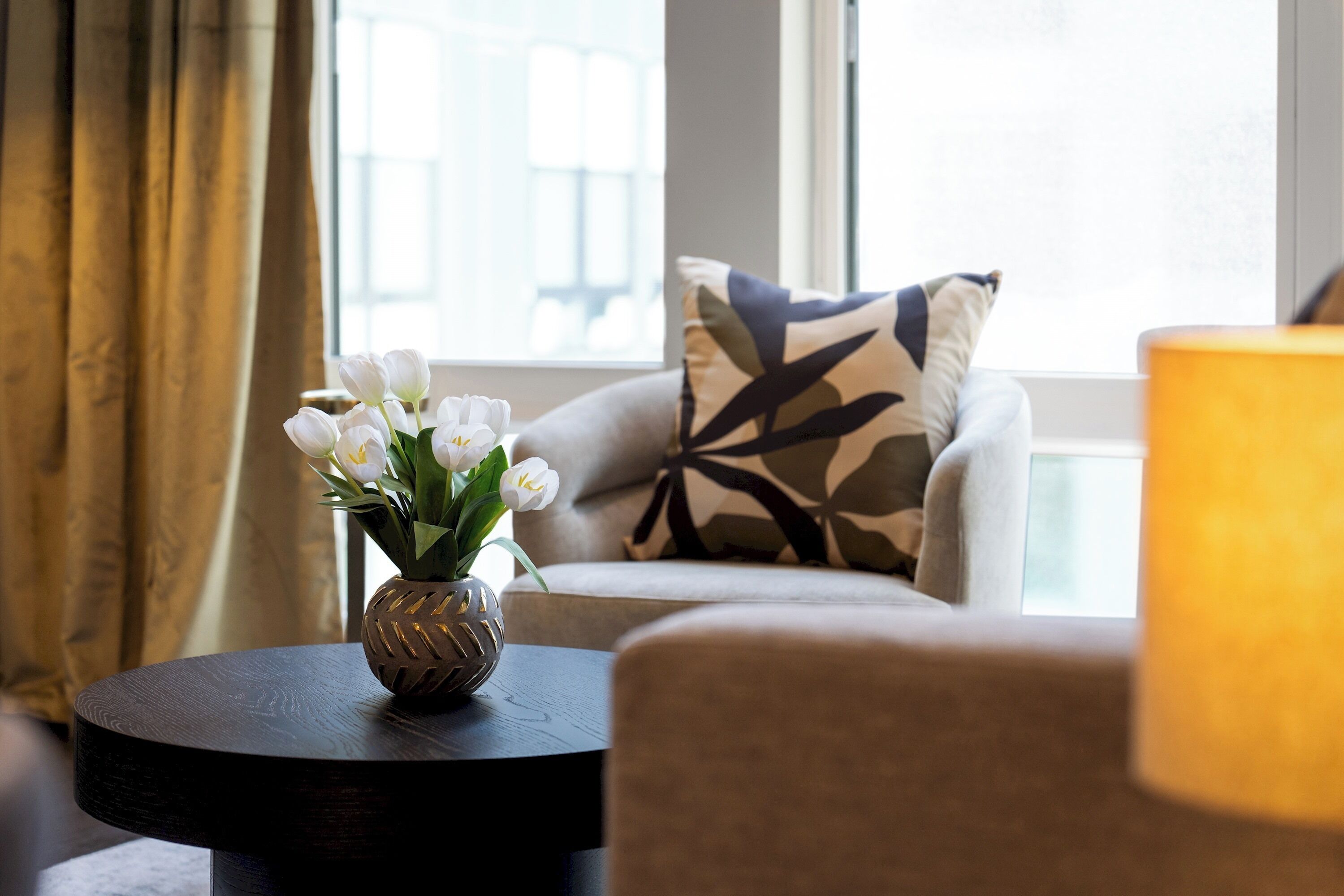 Ein Wohnzimmer mit einer Couch, einem runden Tisch mit einer Vase voller weißer Blumen und einem Fenster mit Vorhängen.