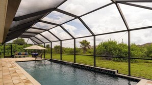 Ein Pool mit Glasveranda, eine Lounge-Bereich mit Stühlen und Sonnenschirm sowie ein Blick auf Grünflächen und Hügel im Hintergrund.