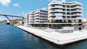 A modern multi-story building with balconies, located near a waterfront with a bridge in the background.