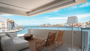A balcony with a sofa, two wicker chairs, a small table, and a floor lamp. The balcony overlooks a waterfront with buildings and a cruise ship.