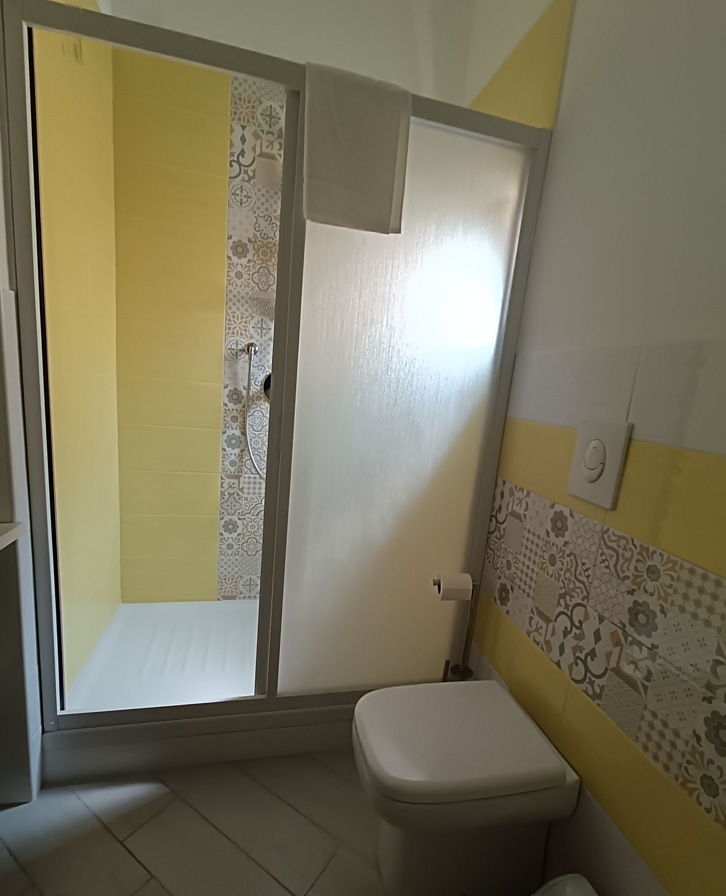 A bathroom with a toilet, a shower with a patterned door, and a tiled wall with a floral design.