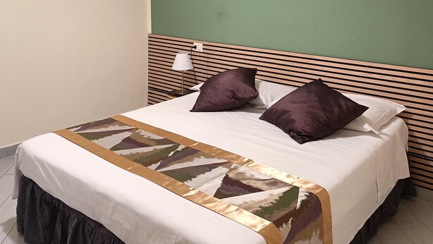 A neatly made bed with a patterned bedspread, two pillows, and a bedside lamp on a wooden headboard. - In the Hospital Zone of Naples. (Napoli)