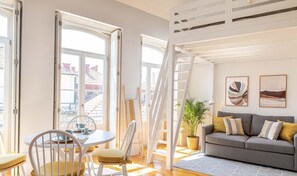 Ein helles Wohnzimmer mit grauem Sofa, einem Esstisch mit Stühlen und großen Fenstern.