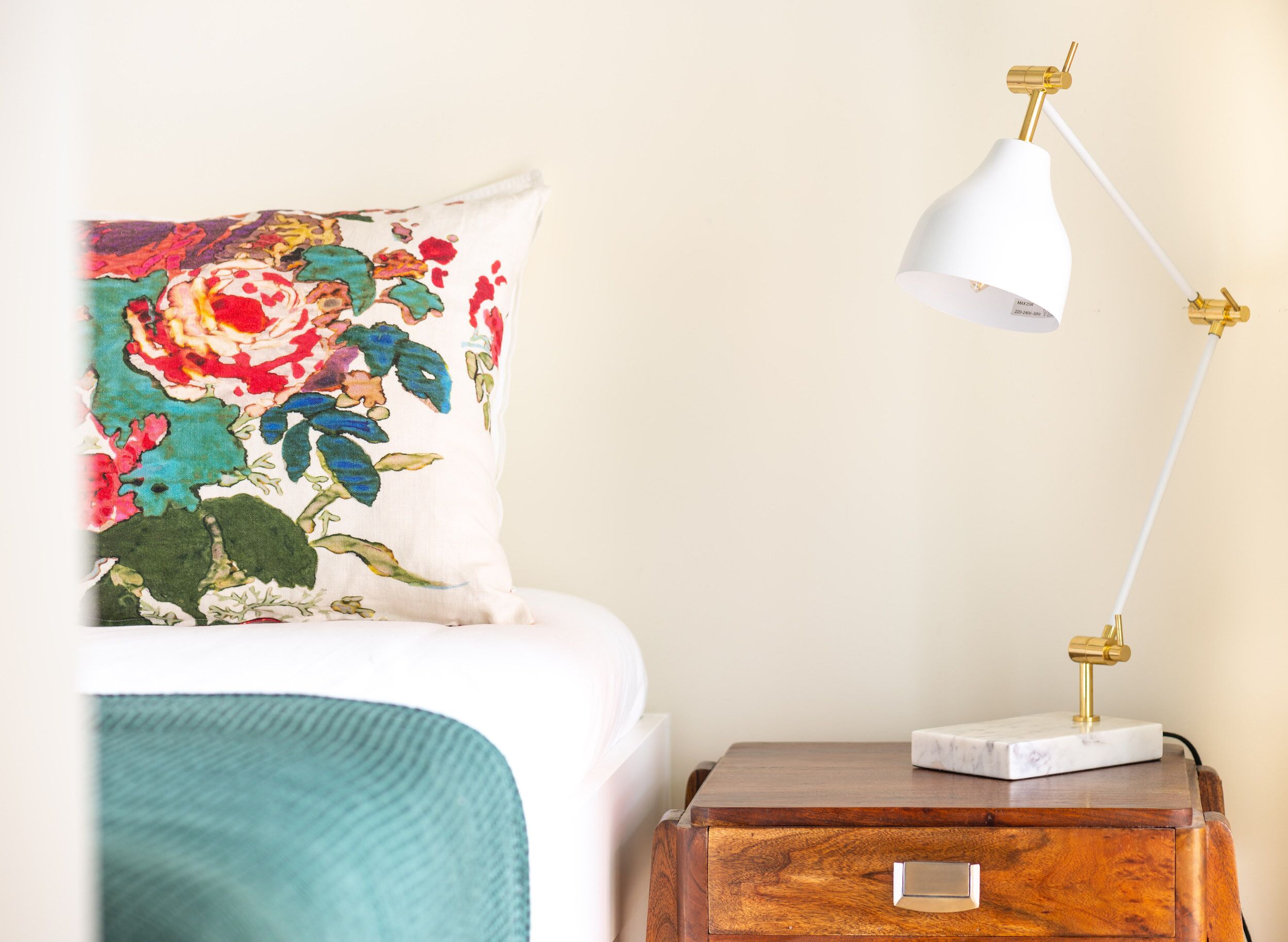 Un lit avec un coussin à motifs floraux, une lampe blanche avec un pied doré et une table de chevet en bois surmontée d’un plateau en marbre.