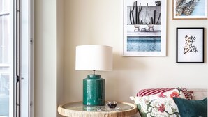 A cozy living room with a wicker side table, a grey ottoman, a sofa with floral cushions, and framed pictures on the wall. - Foz003 · Light-filled Studio With Courtyard Garden (Porto)