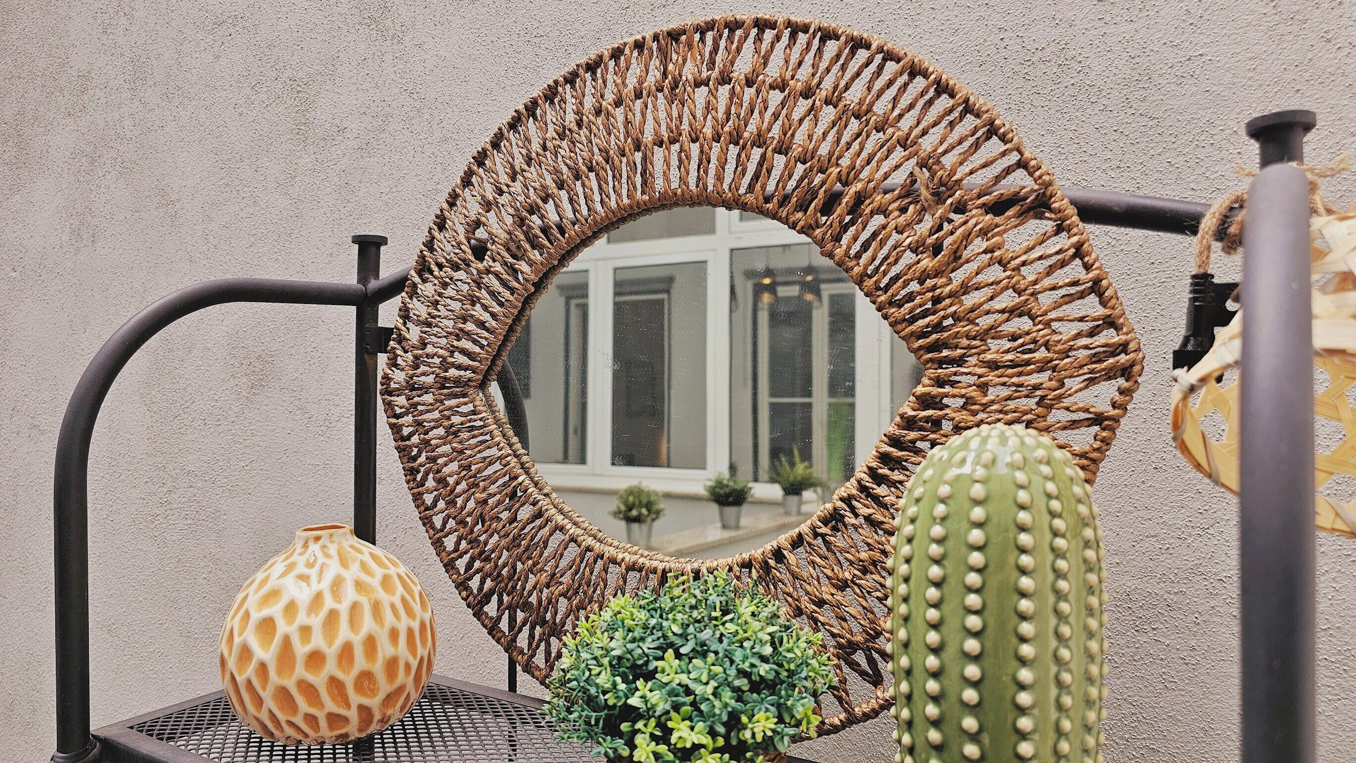 Un espejo redondo tejido con marco metálico, un cactus y un florero texturizado sobre una mesa metálica frente a una pared texturizada.