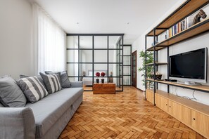 Una sala de estar moderna con un sofá gris, suelo de madera con diseño espiga, un televisor sobre un mueble de madera y una estantería con libros y objetos decorativos.