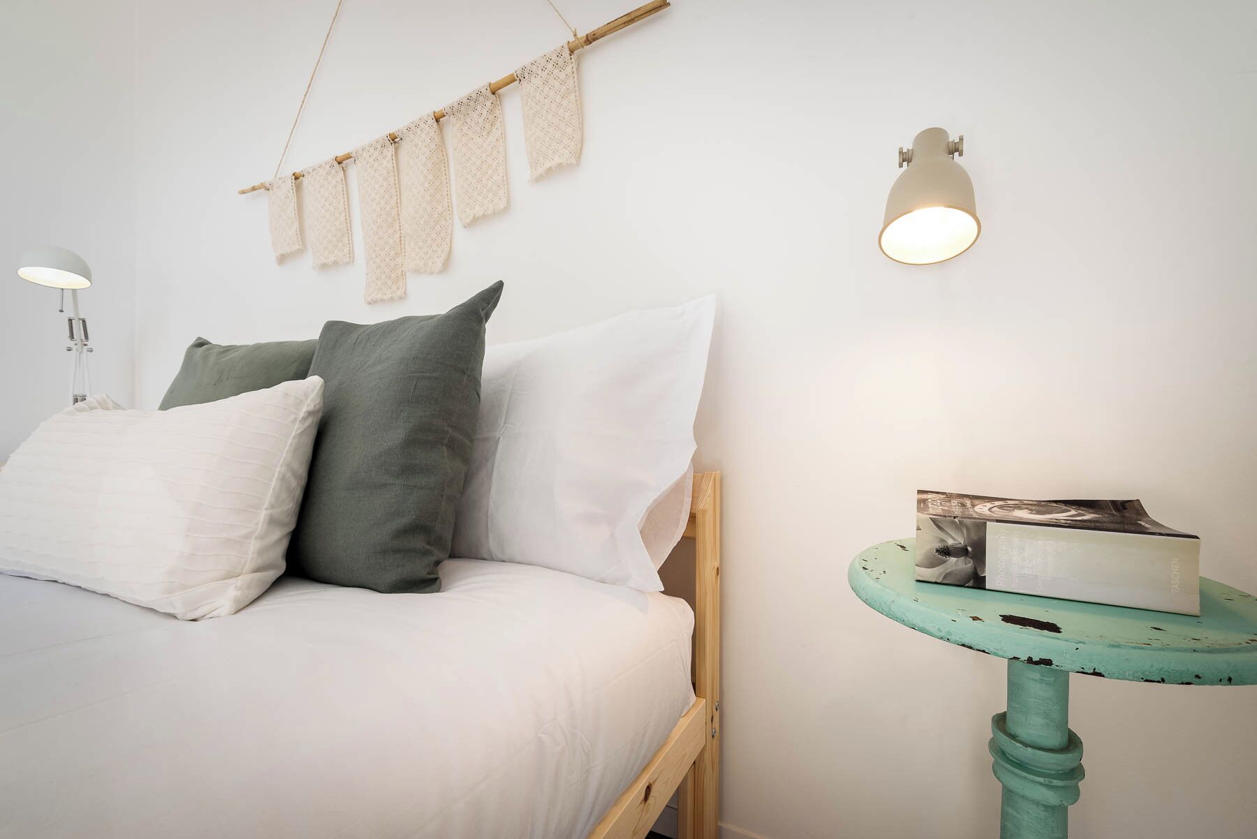 A bedroom with a bed, pillows, a wall-mounted lamp, a small table with a box, and a string of fabric hanging on the wall.