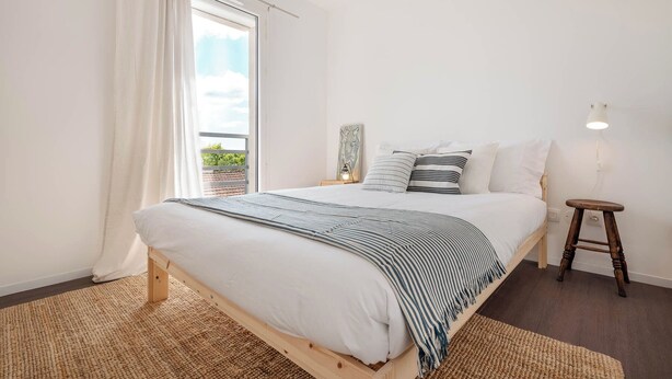 Une chambre à coucher avec un lit, une table de chevet, une lampe, une fenêtre avec des rideaux et une vue sur la verdure à l’extérieur.