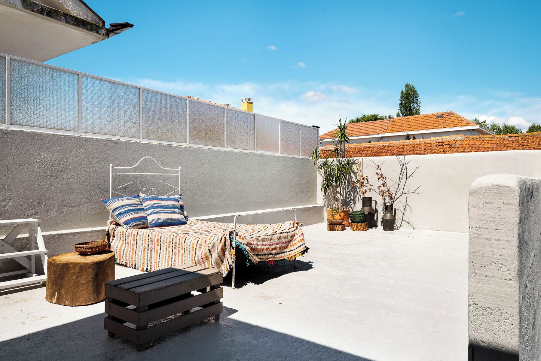 Un espacio exterior privado y pequeño con una cama, un taburete de madera y plantas en macetas.