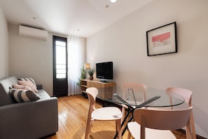 Una sala moderna con un sofá gris, una mesa de comedor de vidrio con sillas, un televisor sobre un mueble de madera y un cuadro enmarcado en la pared.