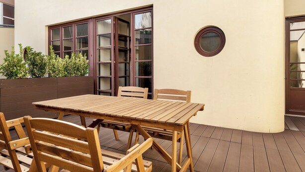 An outdoor patio with a wooden table and chairs, a sliding glass door, and a small round window on a building.
