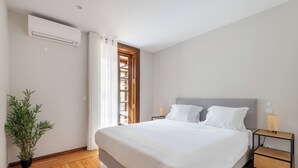 A bedroom with a large bed, a potted plant, a nightstand, a lamp, and a window with curtains.