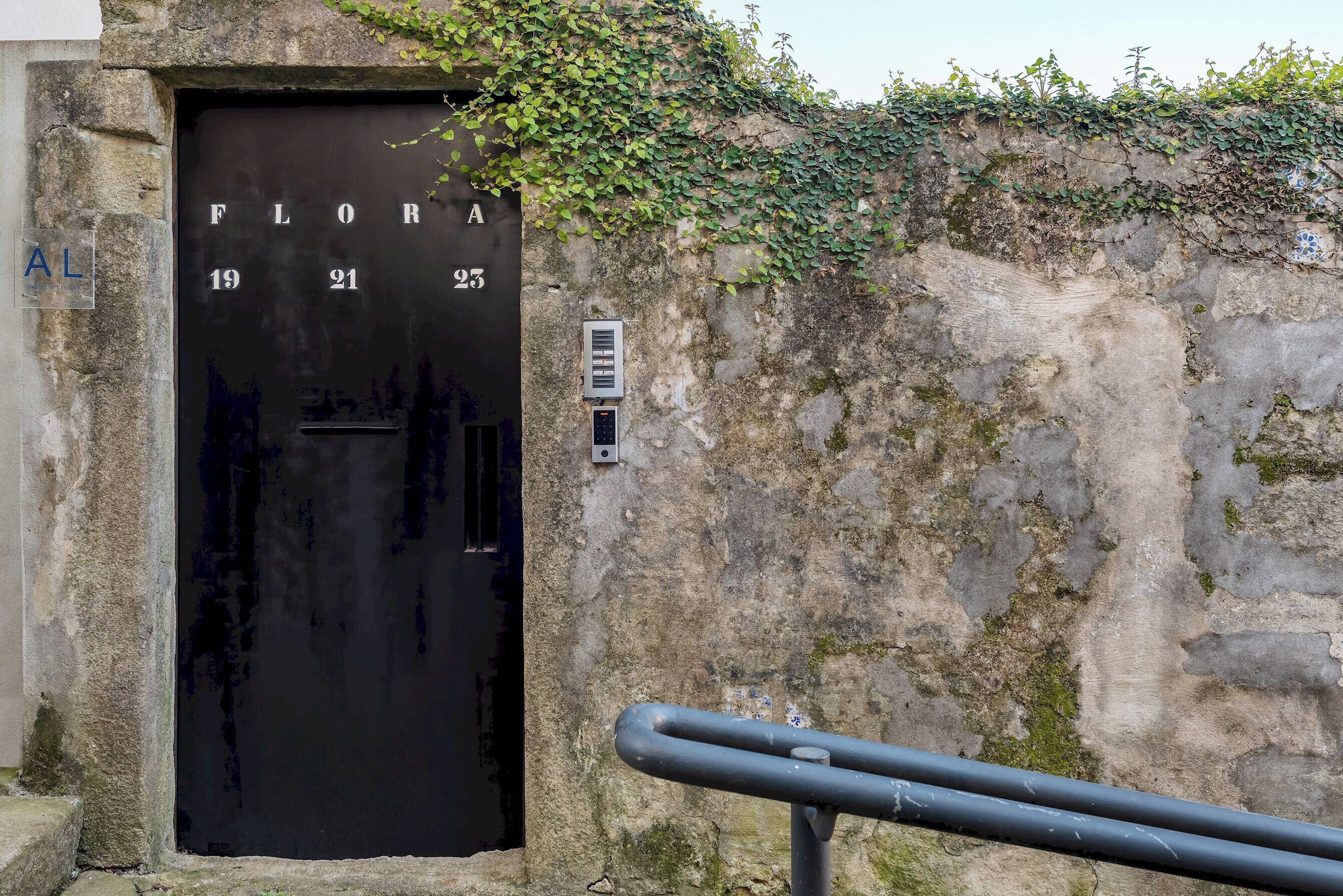 Una puerta negra con la inscripción "FLORA 19 21 23" y un pequeño buzón en la pared.