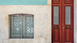 A building facade with a window featuring a metal grille and a red door with glass panels and a brass handle. - Markes · 🪴 Lovely 1BR Home w/ Sunny Backyard (Porto)