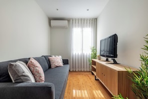 A modern living room with a grey sofa, a wooden TV stand, and a large window with sheer curtains. - Rivol5 · Unique Flat Very Central @rivoli (Porto)