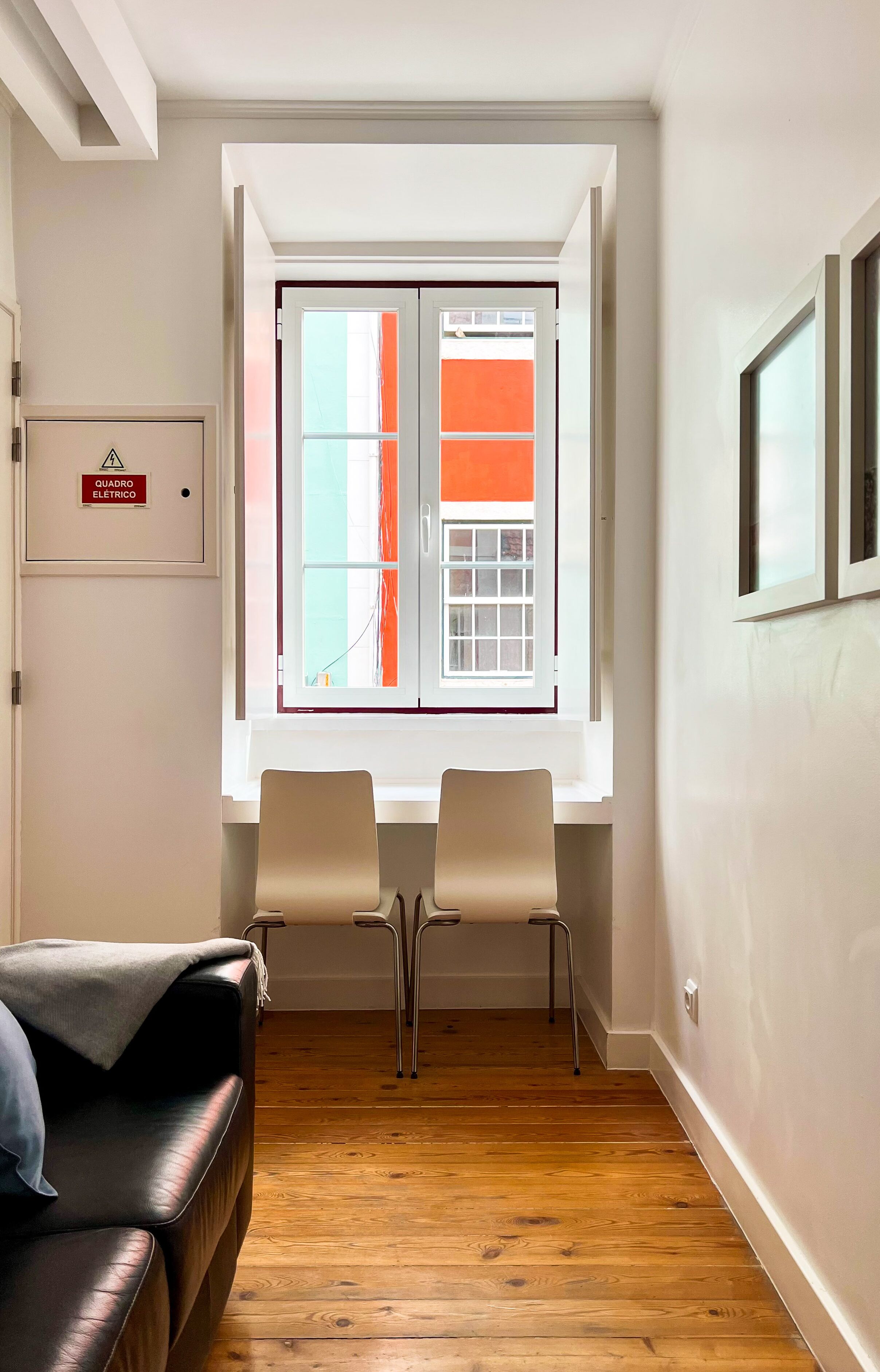 A room with a wooden floor, a black leather sofa, two chairs, a window with orange curtains, and a sign indicating an electrical hazard.