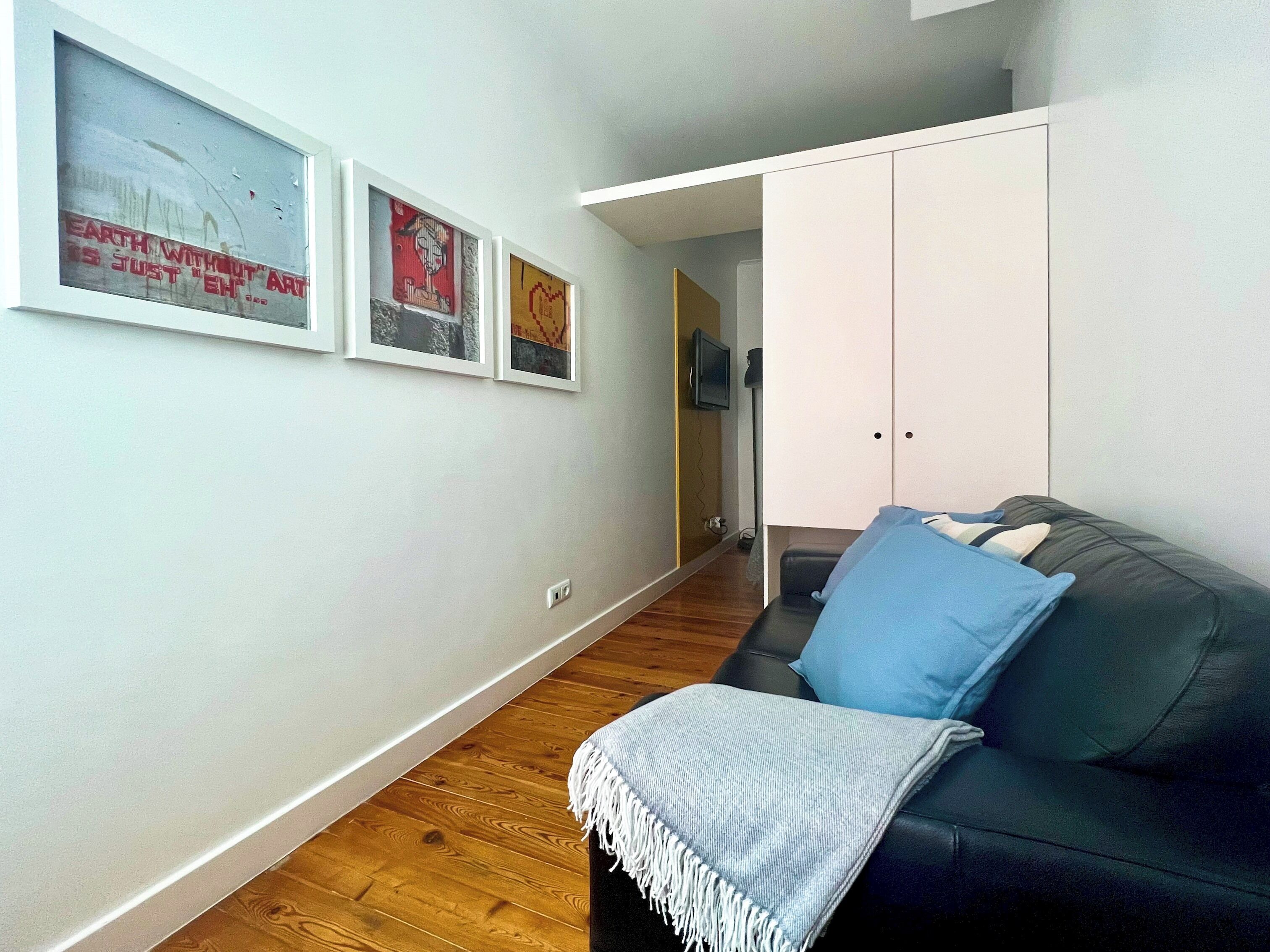 Un salon moderne avec un canapé en cuir noir, un coussin bleu et un plaid gris. Trois œuvres d’art encadrées ornent le mur, et une armoire blanche est visible au fond.