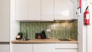 Una cocina con gabinetes blancos, un salpicadero de azulejos verdes, un lavamanos de acero inoxidable, una cafetera y un extintor de incendios colgado en la pared.