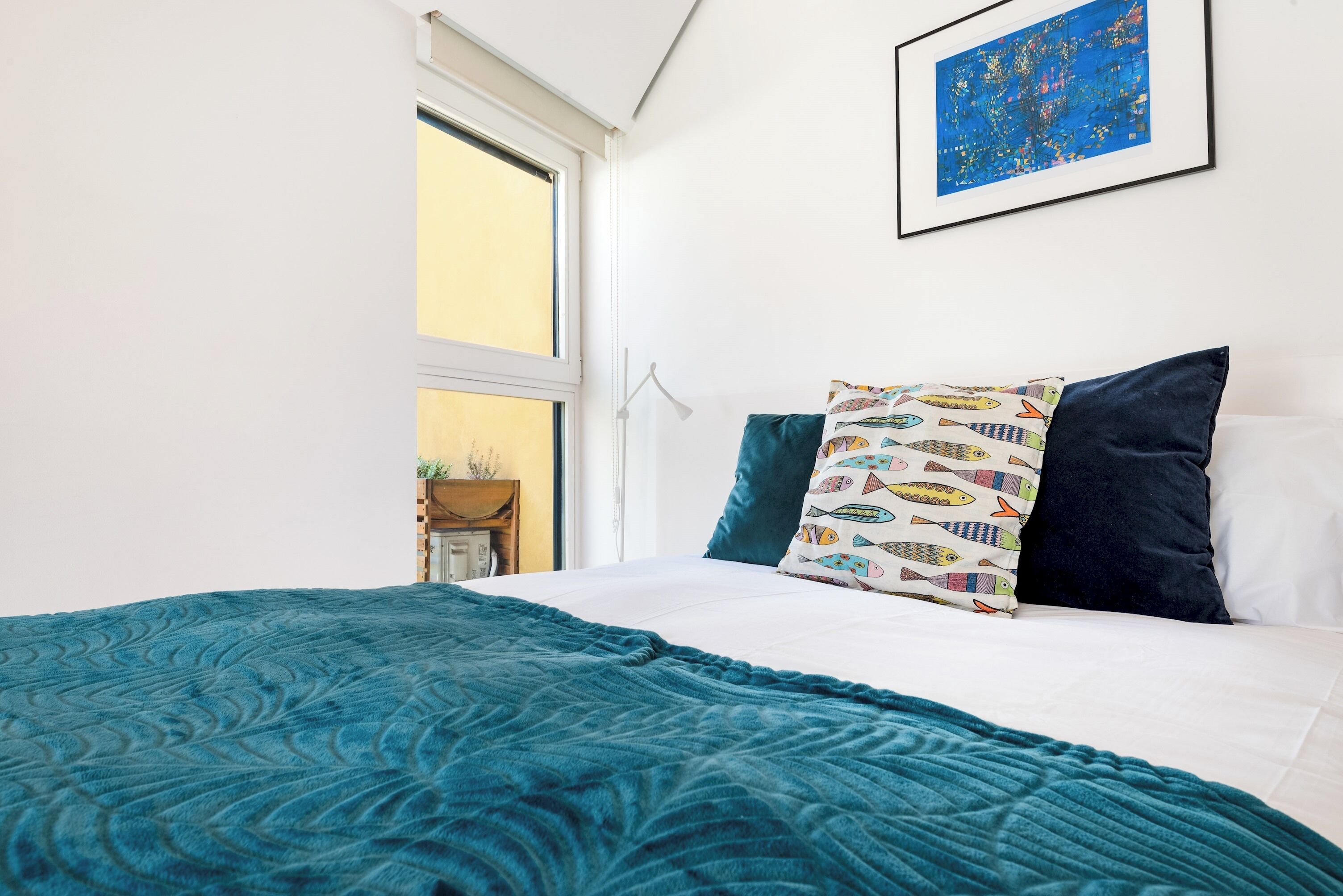 Une chambre à coucher avec un lit, une couverture turquoise, des coussins à motifs, une œuvre d’art encadrée et une fenêtre donnant sur un paysage.