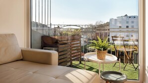 Un balcone con un divano, un tavolino e sedie, affacciato su una zona verde e edifici.