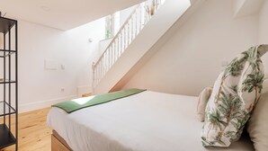 Une chambre avec un lit, un plaid vert, un oreiller blanc à motifs tropicaux, une étagère en métal noir et un escalier avec une rampe blanche.