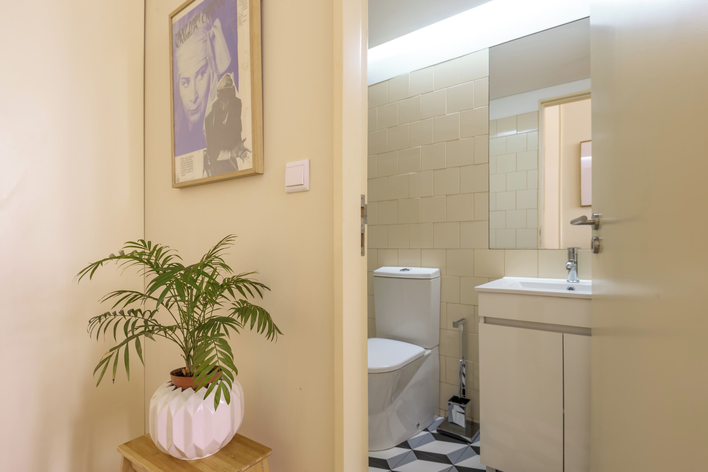 A bathroom with a toilet, sink, and mirror. There is a potted plant on a small table next to the wall.
