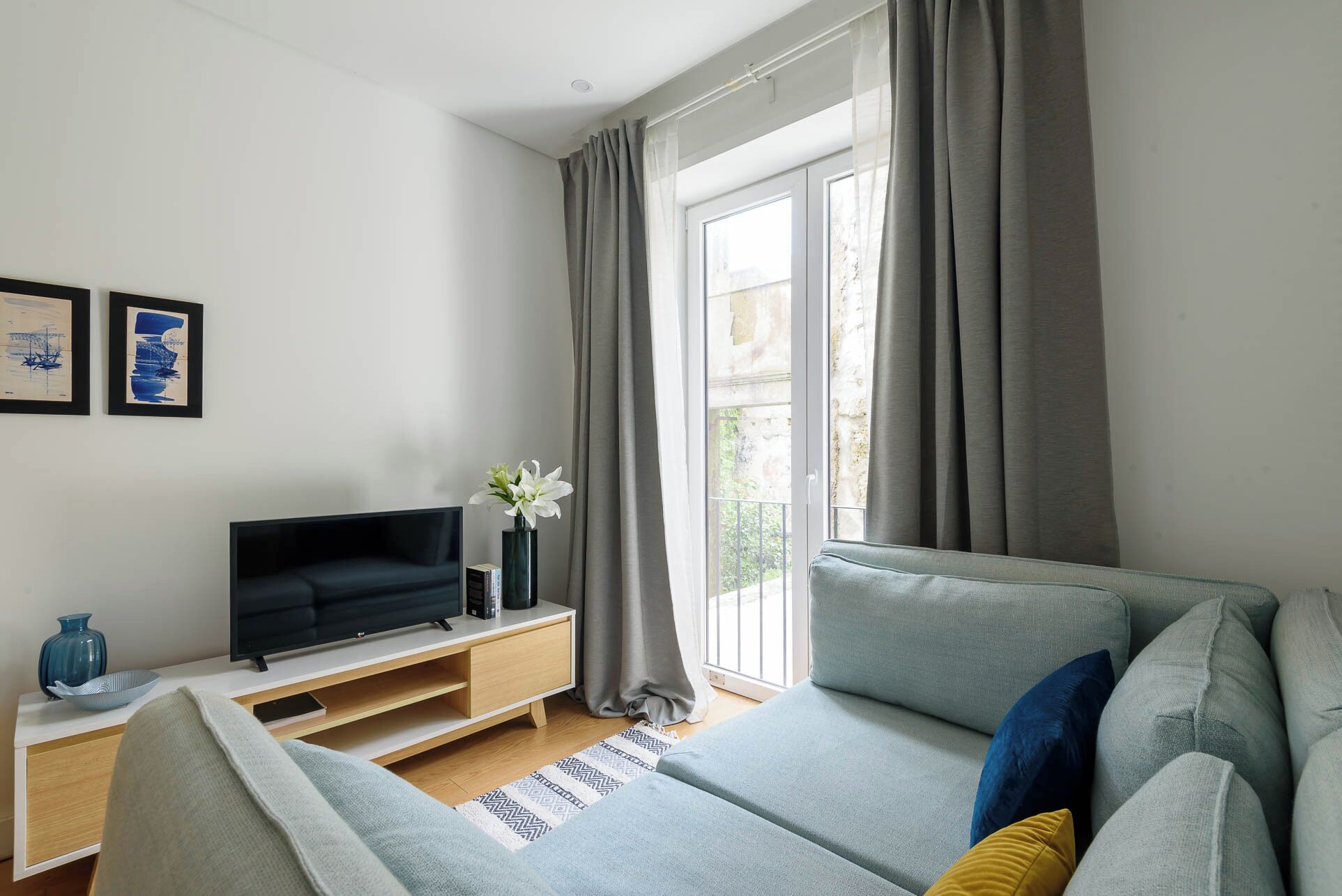 Ett modernt vardagsrum med en grå soffa, en platt-TV på ett träställ, en vas med vita blommor och ett fönster med grå gardiner.