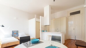 A modern, well-lit room with a round dining table set for two, a sofa, a kitchen area, and a door.