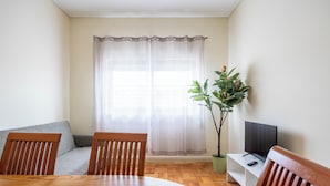 A dining area with a round wooden table, two wooden chairs, a grey sofa, a television on a white stand, and a potted plant. - Marino · 3bed apt Close to Casa da Musica (Porto)