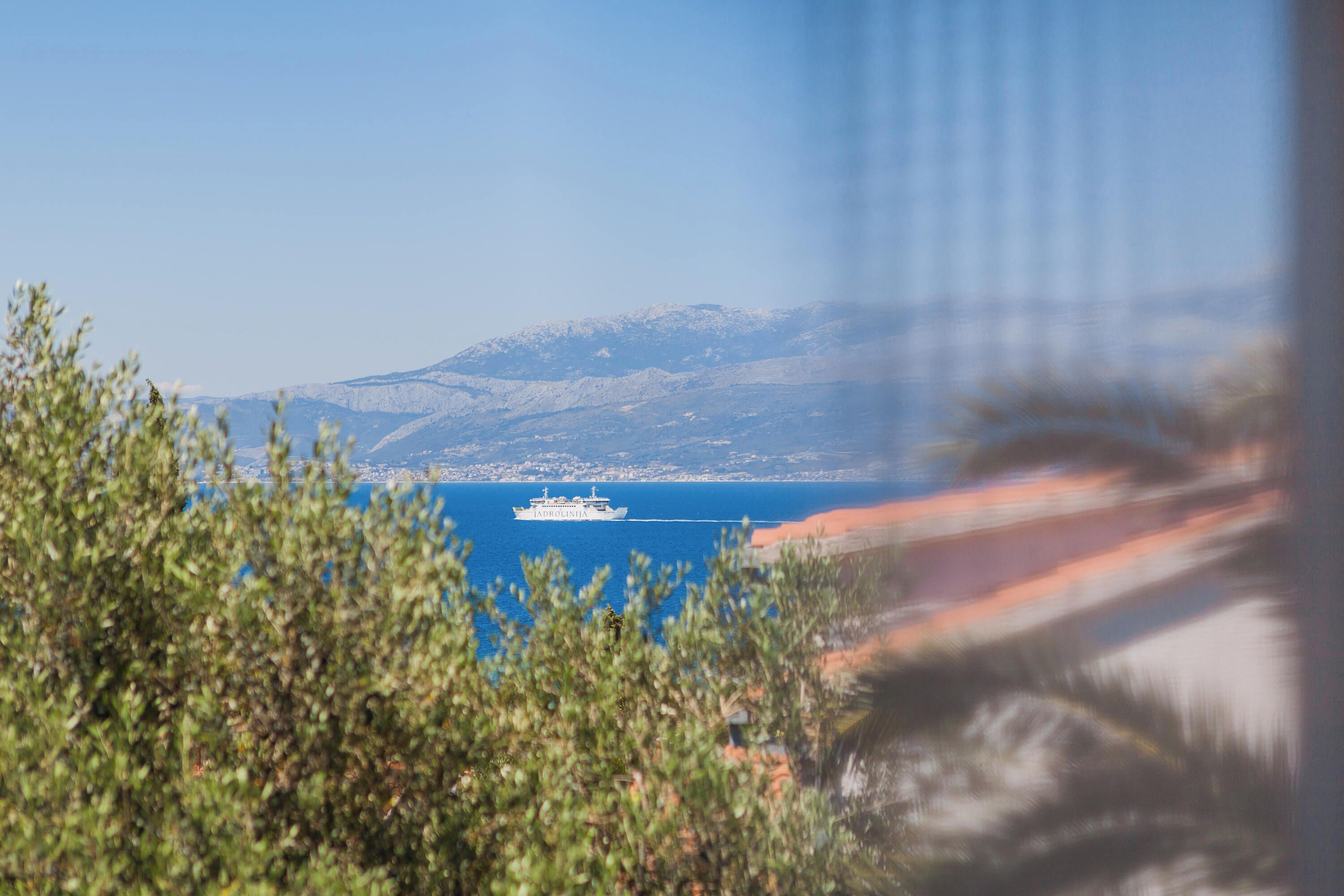 Vista costera con un barco a lo lejos, montañas al fondo y vegetación en primer plano.