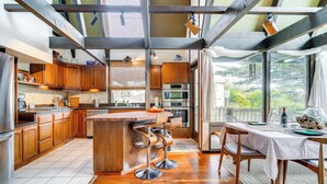 A modern kitchen with wooden cabinets, a central island with bar stools, stainless steel appliances, and large windows offering a view of greenery. - Moss Beach Oceanfront Retreat ~ 20 Mi to San Fran! (Moss Beach)