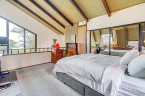 A bedroom with a large bed, a wooden ceiling, a window with a view of trees, and a desk with a lamp.