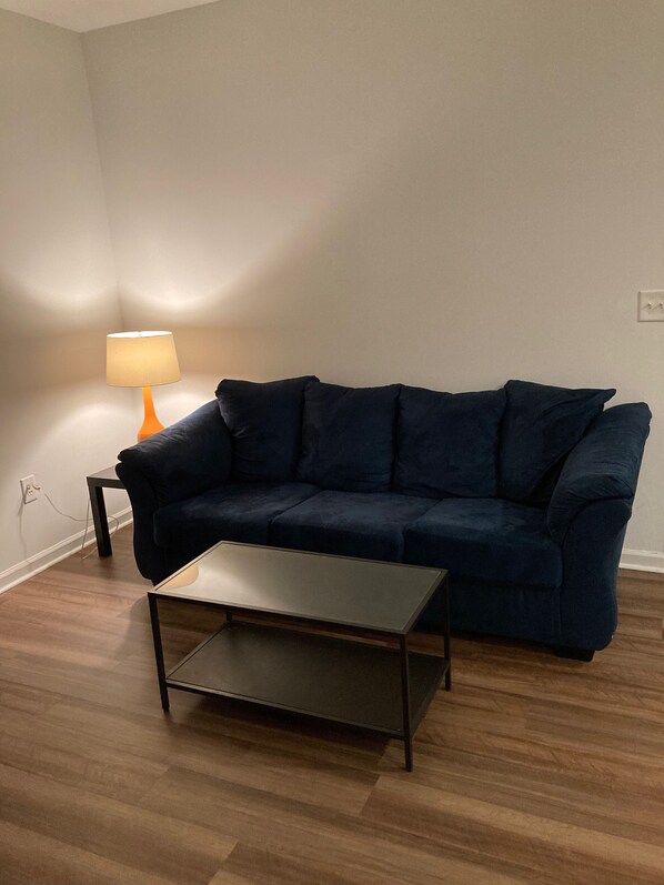 Un sofá azul oscuro, una mesita y una lámpara en una habitación con suelo de madera y paredes blancas.