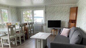 A living room with a grey sofa, a white coffee table, and a TV. There is a dining area with a table and chairs, and a window with a view. - House at the lake, 3 Bedrooms, WIFI, nature (Västra Götalands län)