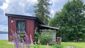 A cabin with a red exterior, a wooden deck, and a garden with various plants and flowers. - House at the lake, 3 Bedrooms, WIFI, nature (Västra Götalands län)