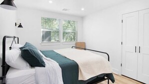 A single bed with a black frame, white bedding, and a green accent pillow. There is a window with blinds, a white wardrobe, and a black floor lamp. - Modern Backyard Casita (Bryan)