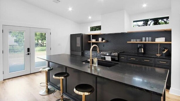 A modern kitchen with a black countertop, stainless steel appliances, and a large island with bar stools. - Modern Backyard Casita (Bryan)
