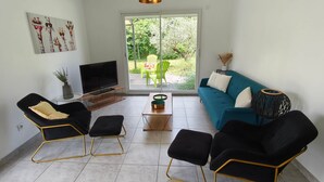 Un salon comprenant un téléviseur, un canapé et des fauteuils, une porte-fenêtre coulissante donnant sur un jardin, et une petite table sur laquelle est posée une plante.