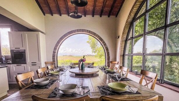A dining area with a large table set for six, surrounded by wooden chairs, with a view of a vineyard through a large arched window. - Villa surrounded by vineyards with private pool near Orvieto (Terni)