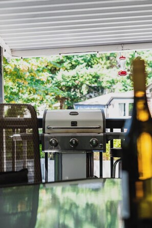 A barbecue grill with controls, a bottle of wine, and a view of trees and a house in the background.
