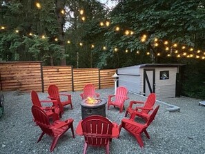 Eine Terrasse mit roten Stühlen, die um eine Feuerstelle angeordnet sind, umgeben von einem Holzzaun und einem Schuppen.