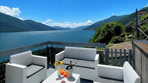 A deck with white wicker furniture and a table with drinks and a bottle, overlooking a lake and mountains. - Rooftop cannobio (San Bartolomeo)