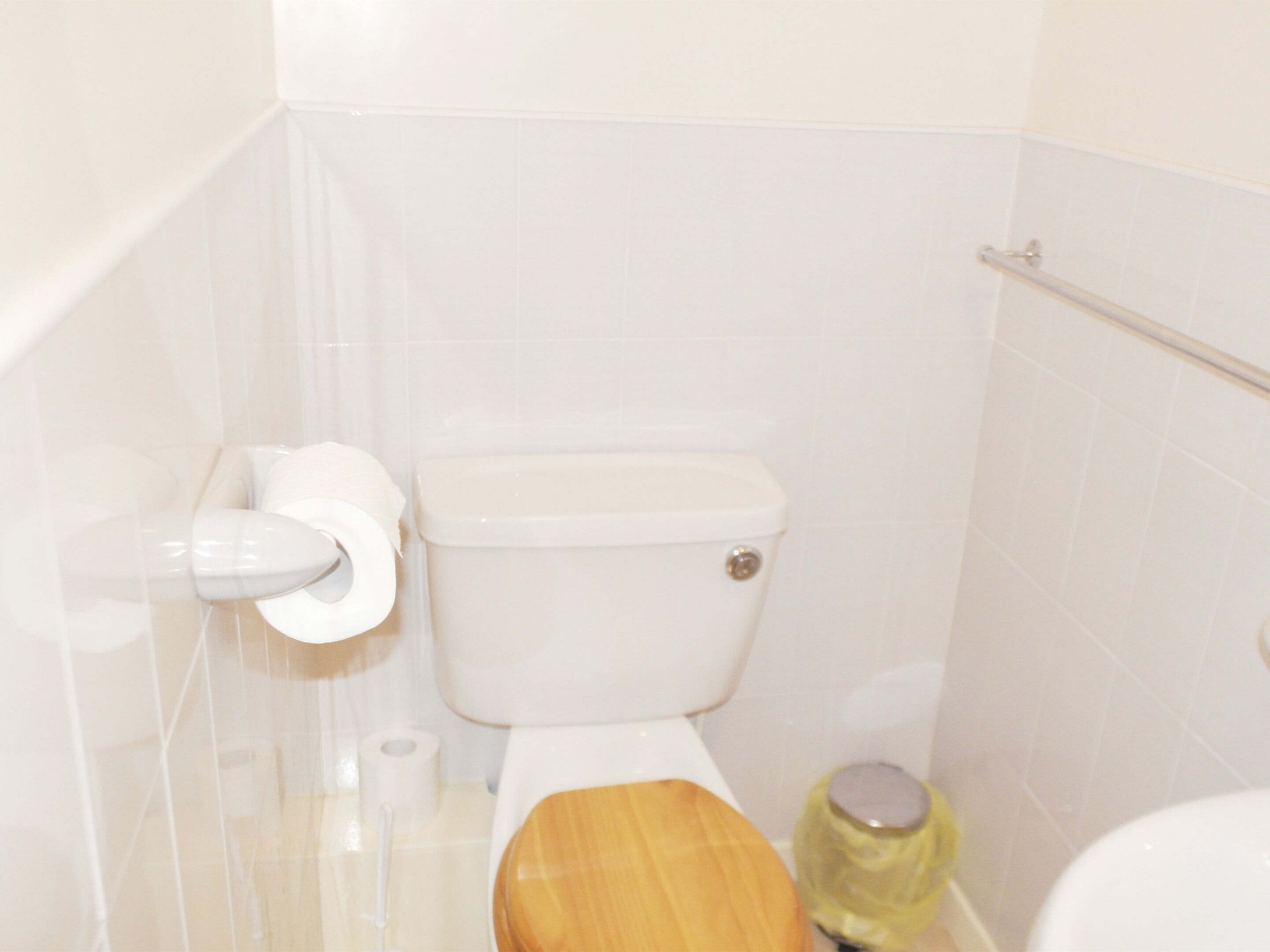 A bathroom with a toilet, a wooden seat, a roll of toilet paper, and a trash bin.